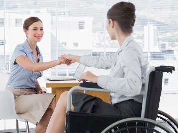 A person shaking hands with a person in a wheelchair. They are in an office in formal clothing.